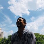 From below ethnic male in striped shirt laughing at joke against cloudy sky on sunny day in city park