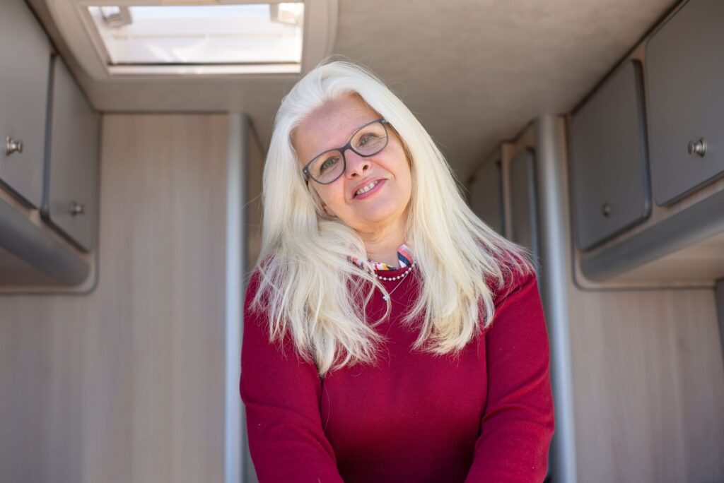 A cheerful woman with glasses and white hair smiling warmly inside a cozy camper van.