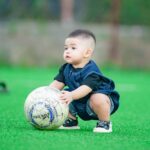 Cute baby playing with a soccer ball on a vibrant green grass field in Hanoi, Vietnam.