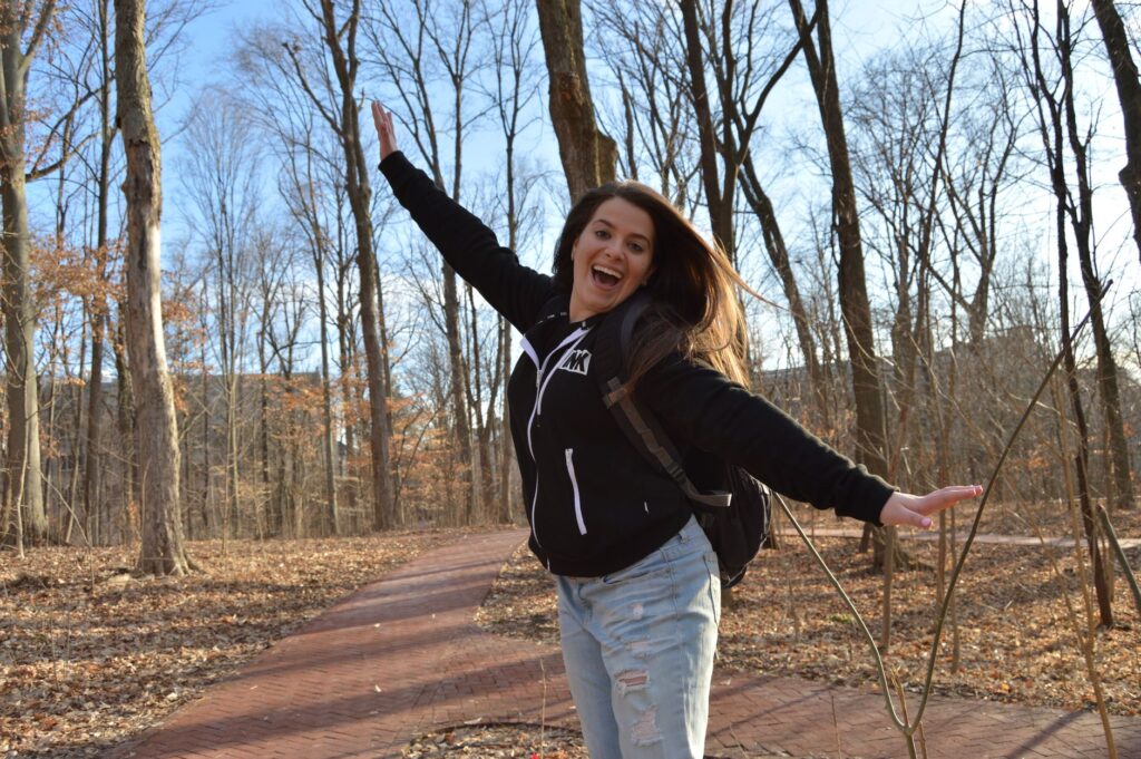 A cheerful young woman in casual attire joyfully posing in Bloomington's Dunn Woods on a sunny winter day.