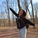 A cheerful young woman in casual attire joyfully posing in Bloomington's Dunn Woods on a sunny winter day.