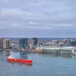 A red cargo ship sails on a calm river against a modern city skyline under a cloudy sky.