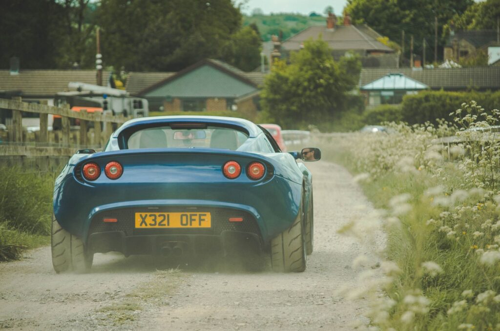 Blue Lotus Elise driving on a dusty road in a quaint small town environment.