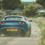 Blue Lotus Elise driving on a dusty road in a quaint small town environment.