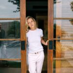 A woman in a white shirt stands by a wooden sliding door, with a reflection visible.