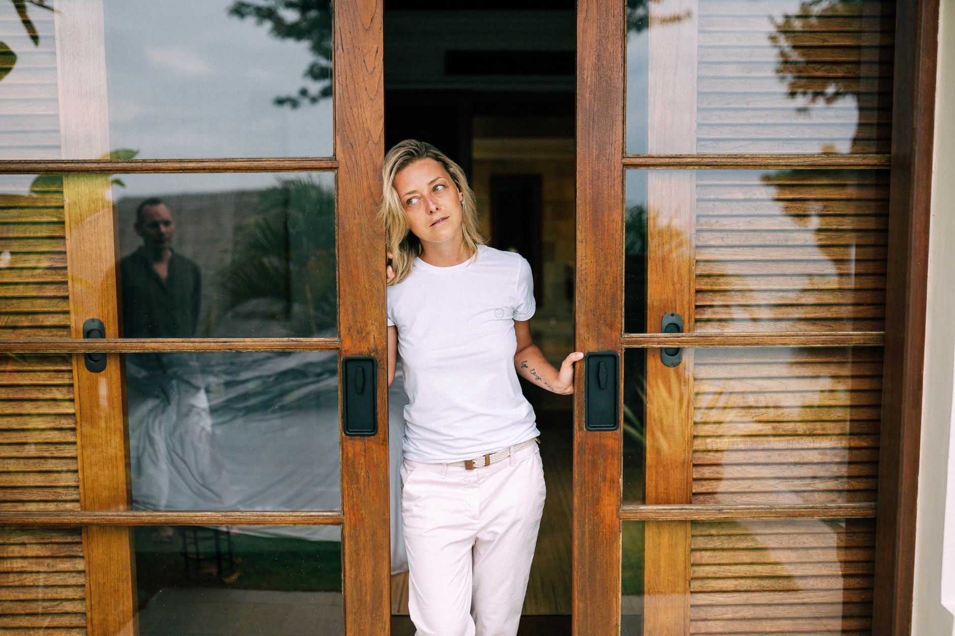 A woman in a white shirt stands by a wooden sliding door, with a reflection visible.