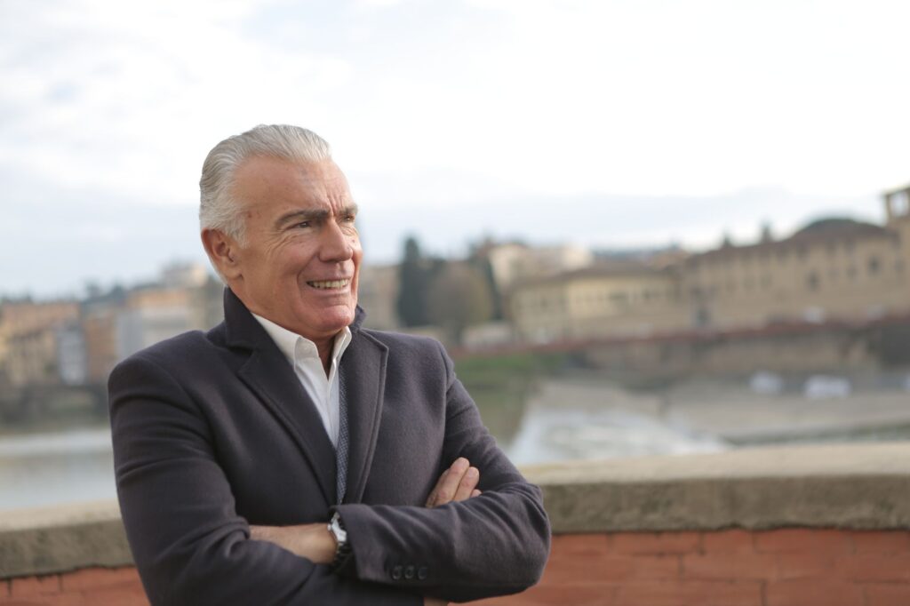 Senior man with arms crossed smiling outdoors with a scenic view in the background.