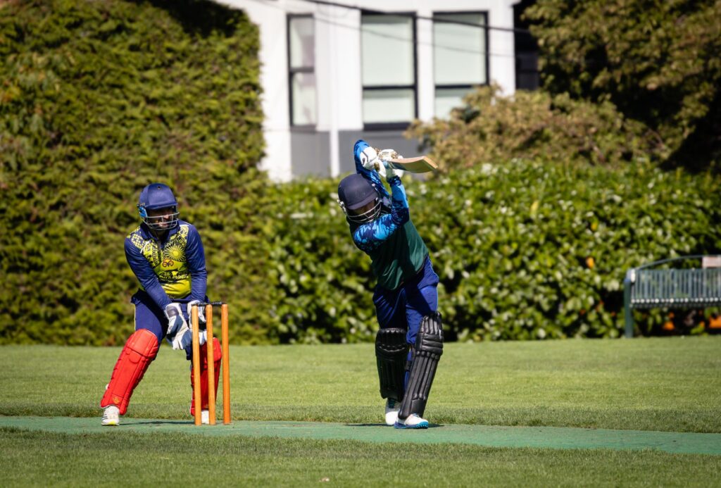 Action shot of a cricket batsman playing a shot while the wicketkeeper looks on during a match outdoors.