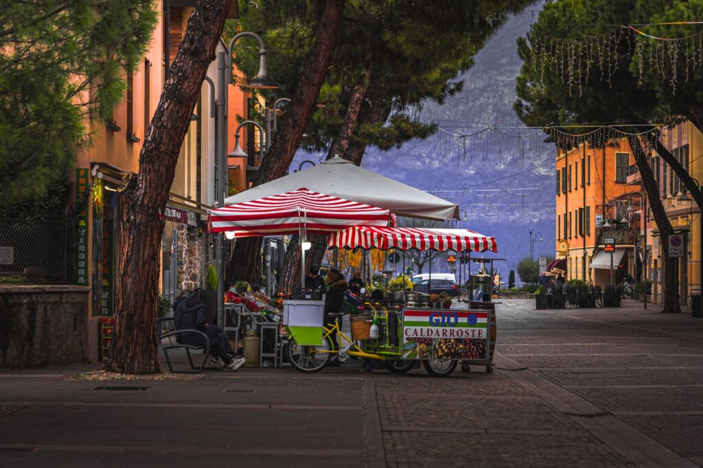 A picturesque street scene featuring colorful outdoor market stalls under pine trees on a quaint cobblestone road.