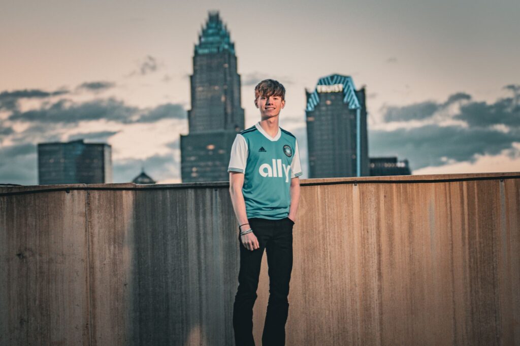 A young adult poses in front of a city skyline during sunset, wearing a sports jersey.