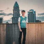 A young adult poses in front of a city skyline during sunset, wearing a sports jersey.
