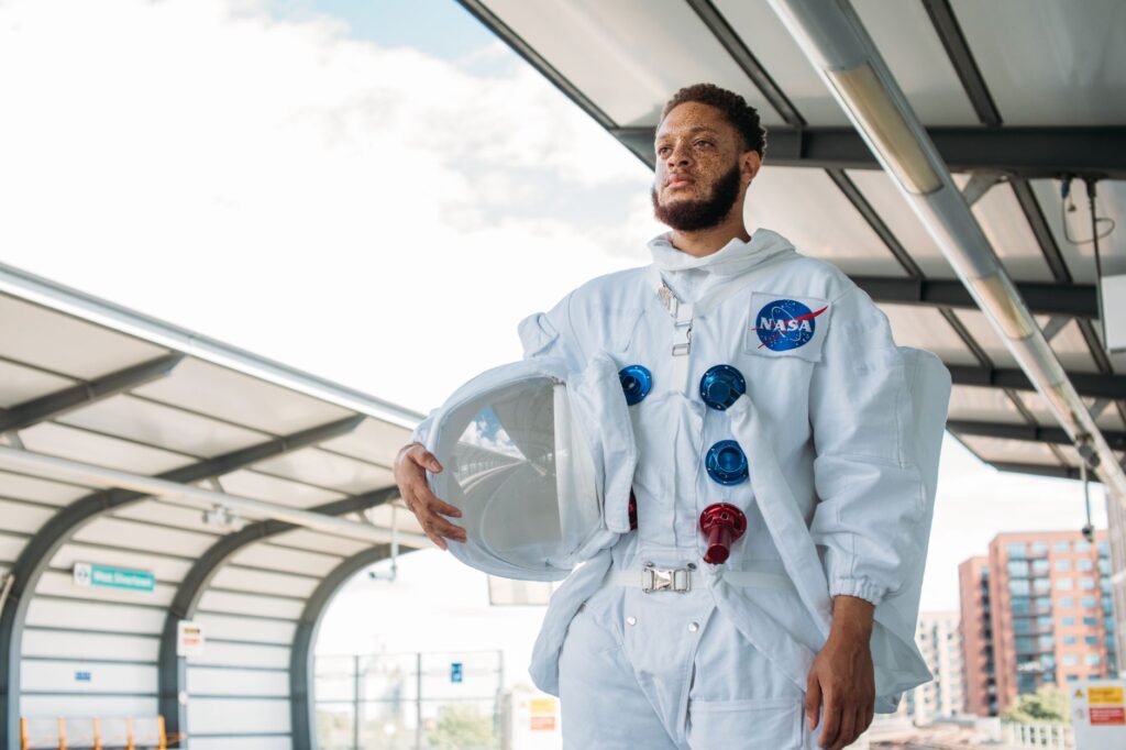 Man in a NASA space suit standing at a city train station, holding helmet.