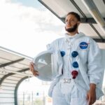 Man in a NASA space suit standing at a city train station, holding helmet.