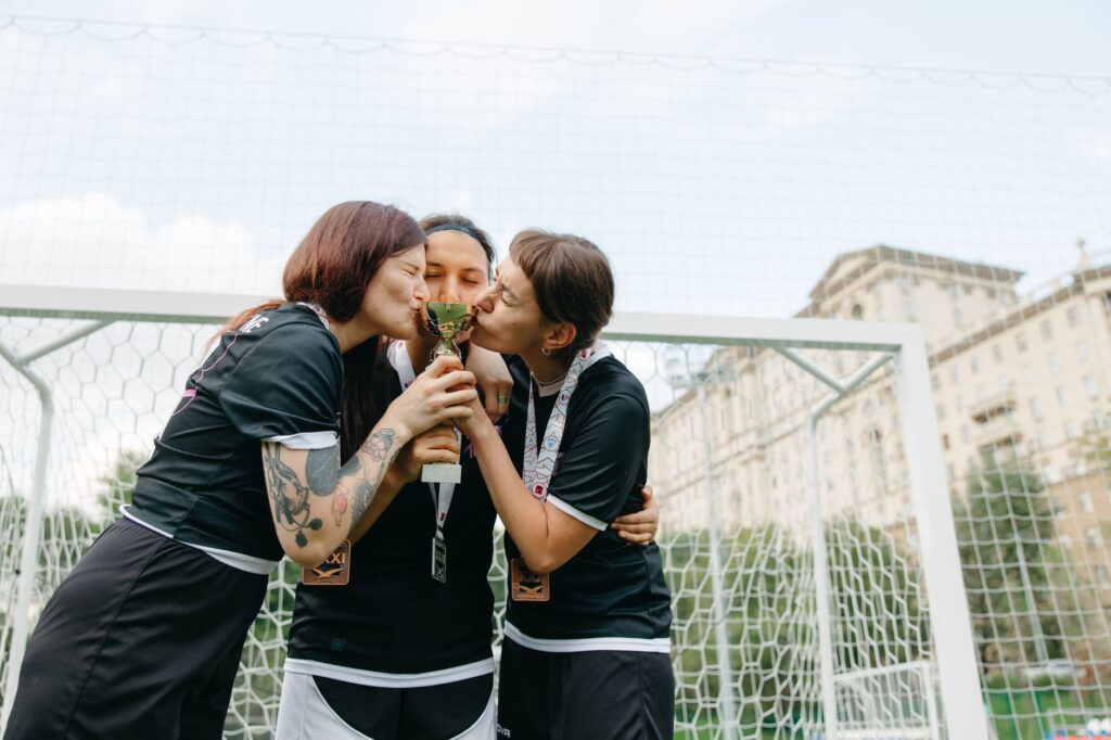 Three female soccer players celebrate winning with a trophy outdoors.