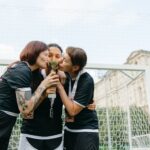 Three female soccer players celebrate winning with a trophy outdoors.