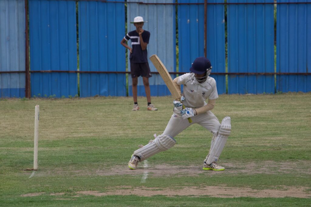 Cricket player batting on a grass field in Gahunje, India, showcasing dynamic sports action.