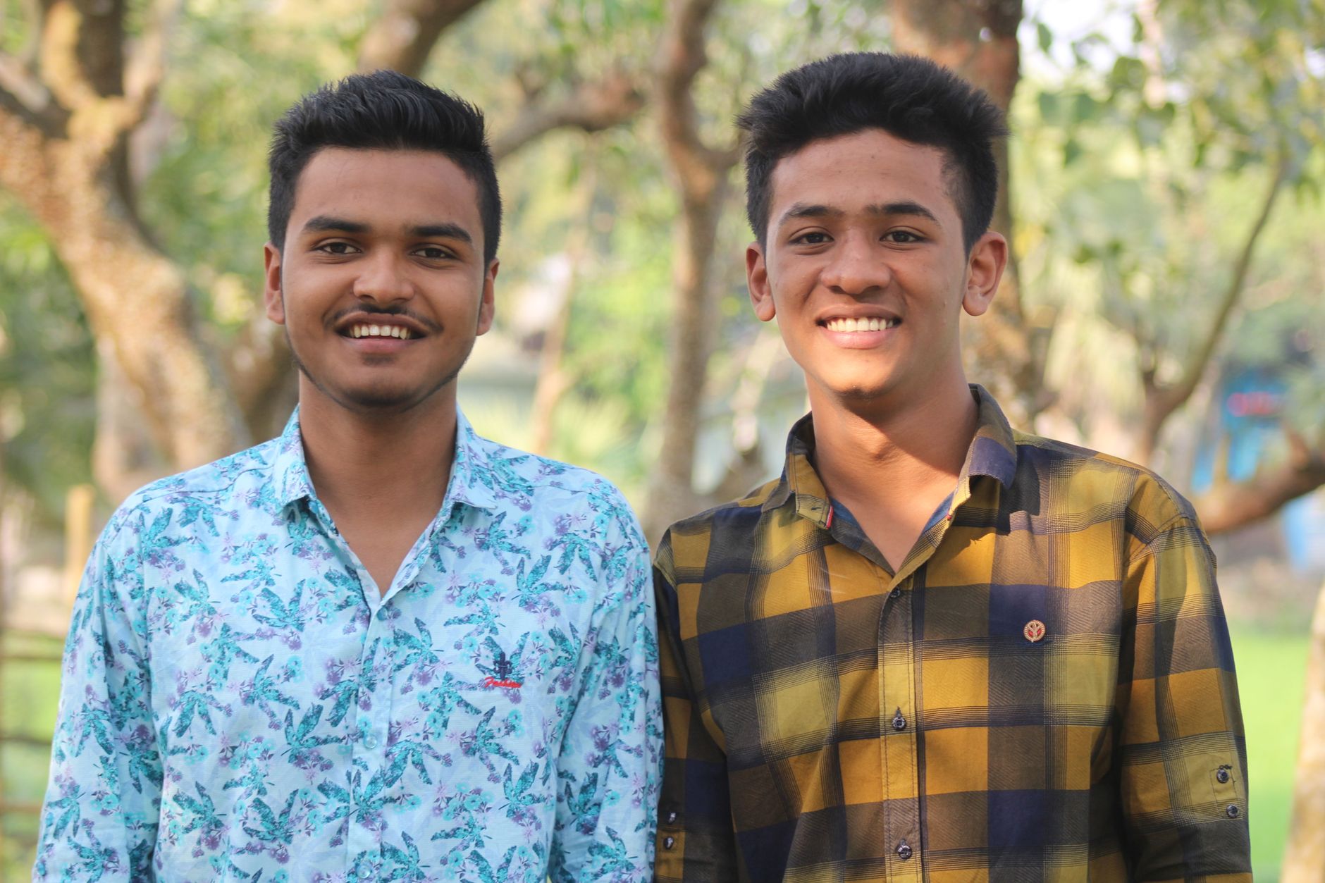 Two young men smiling outdoors in a sunny park, enjoying a summer day.