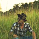 A man wearing sunglasses and a hat sits in a lush green field.