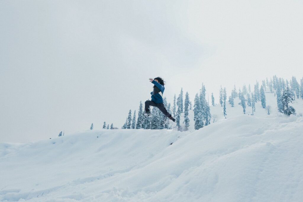 A man leaps across a snow-covered hillside against a backdrop of trees in Gulmarg.