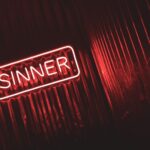 Bright red neon sign displaying the word 'SINNER' against a dark, moody background.