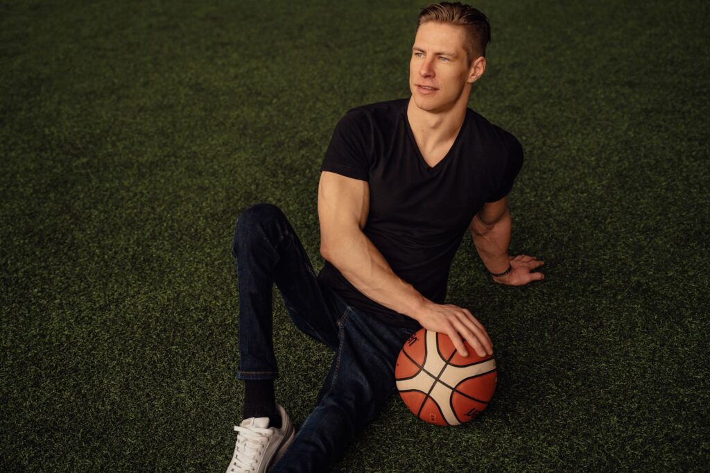 A man sits on artificial grass holding a basketball, dressed in casual attire and looking thoughtfully away.