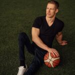 A man sits on artificial grass holding a basketball, dressed in casual attire and looking thoughtfully away.