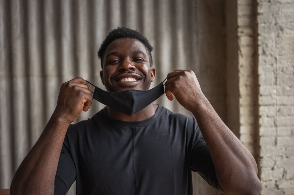 Happy African American male in t shirt with textile mask looking at camera against brick wall