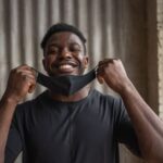 Happy African American male in t shirt with textile mask looking at camera against brick wall