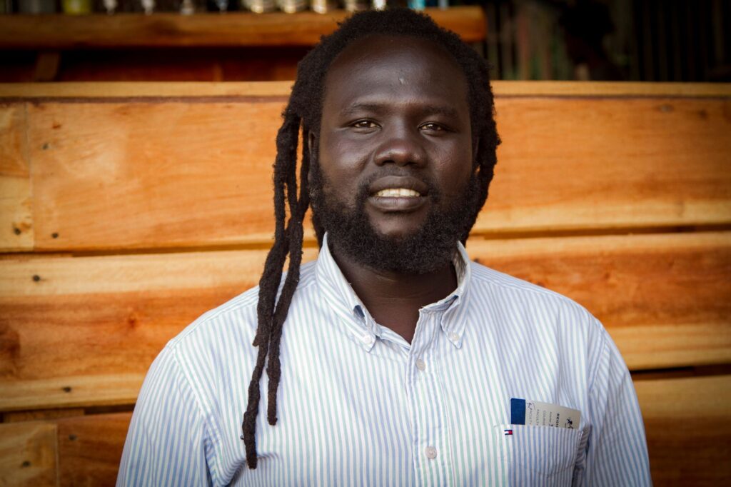 Close-up of a smiling man with dreadlocks and beard against a wooden background.