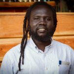 Close-up of a smiling man with dreadlocks and beard against a wooden background.