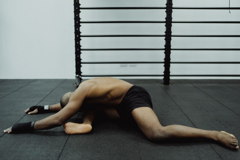 Shirtless man stretches while sitting on gym floor, showcasing flexibility.