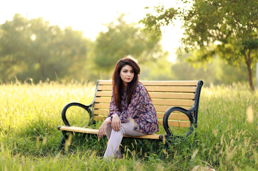 A woman sits thoughtfully on a park bench in a sunny grassy field, embracing nature's tranquility.