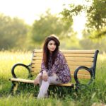 A woman sits thoughtfully on a park bench in a sunny grassy field, embracing nature's tranquility.
