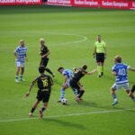 Soccer players in action during a competitive match under bright sunlight.