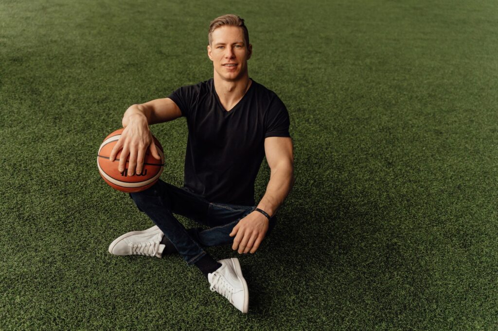 Muscular man sitting on grass holding basketball, wearing casual attire.
