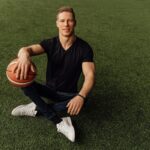 Muscular man sitting on grass holding basketball, wearing casual attire.