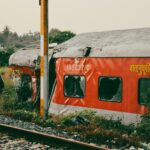 An abandoned and damaged train car rests beside tracks in a lush rural area.