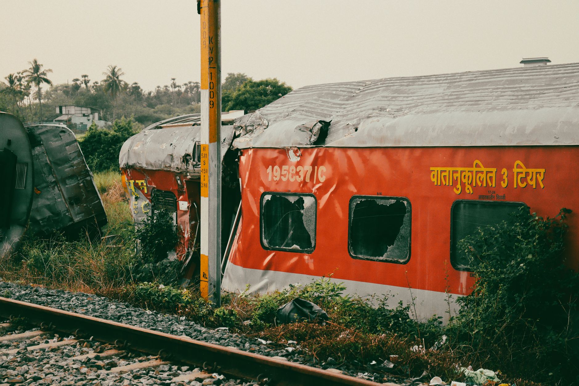 An abandoned and damaged train car rests beside tracks in a lush rural area.