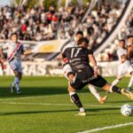 A dynamic football match with players in motion on a sunny day, witnessed by a packed stadium.