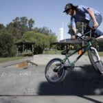 BMX rider performing mid-air trick at a sunny skatepark with green trees and graffiti.
