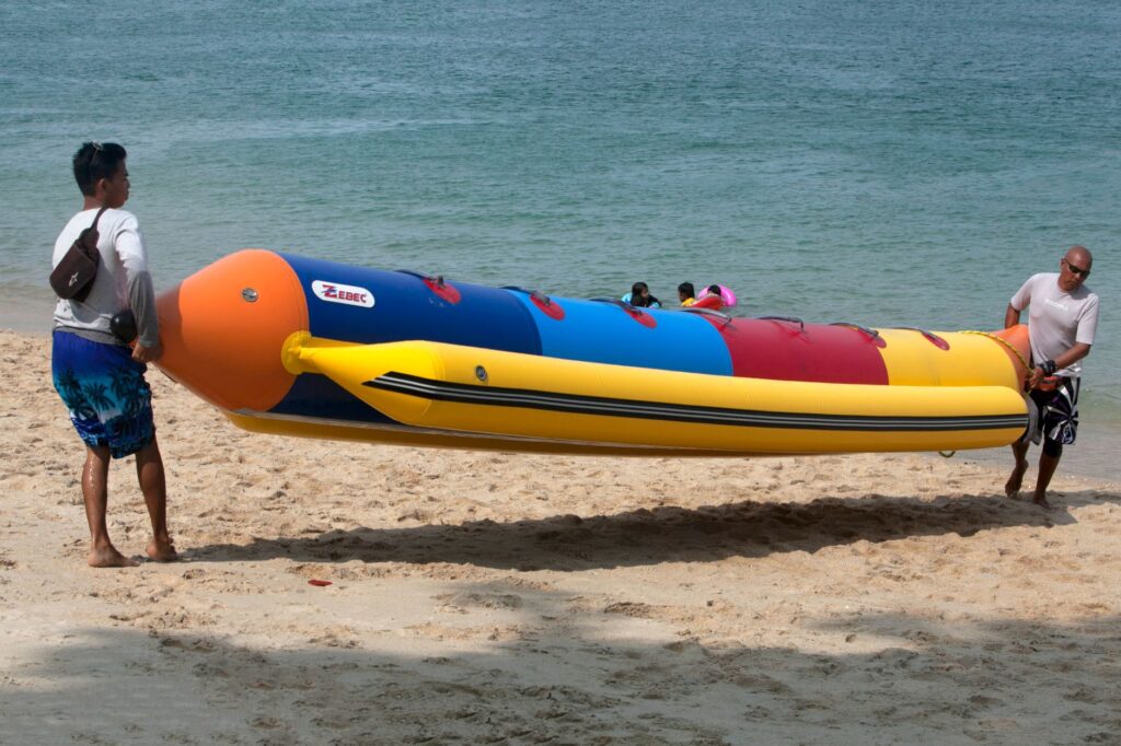 Group carrying a colorful inflatable banana boat along a sandy beach by the sea.