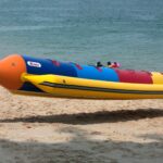 Group carrying a colorful inflatable banana boat along a sandy beach by the sea.