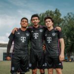 Three young adult soccer players in matching jerseys standing arm in arm outdoors.