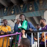 A lively public gathering with diverse participants singing and speaking under an urban bridge setting.