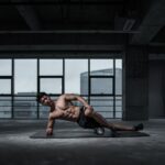 Muscular man performing yoga stretch indoors on a mat with urban window view in the background.