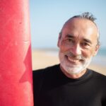 Elderly man with a surfboard enjoying a sunny day at a Portuguese beach.