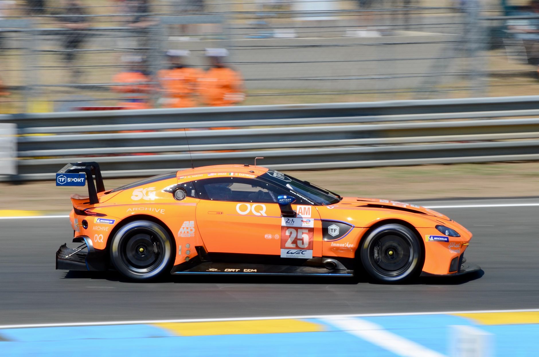 A bright orange Aston Martin on the Le Mans circuit, captured during a motorsport endurance race.