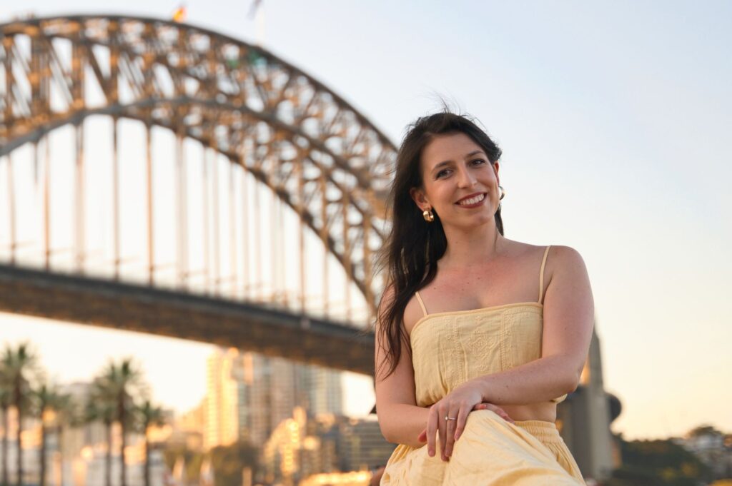 A cheerful woman poses in front of the iconic Sydney Harbour Bridge during sunset, creating a memorable moment.
