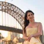 A cheerful woman poses in front of the iconic Sydney Harbour Bridge during sunset, creating a memorable moment.