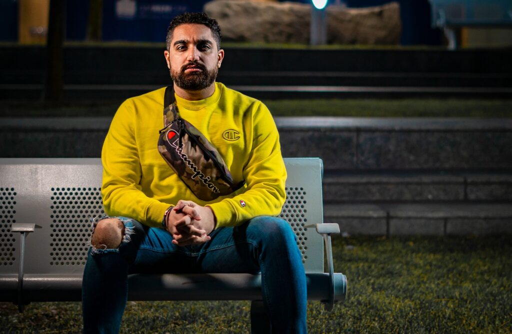 A man in a yellow sweater and jeans sits on an outdoor bench at night.
